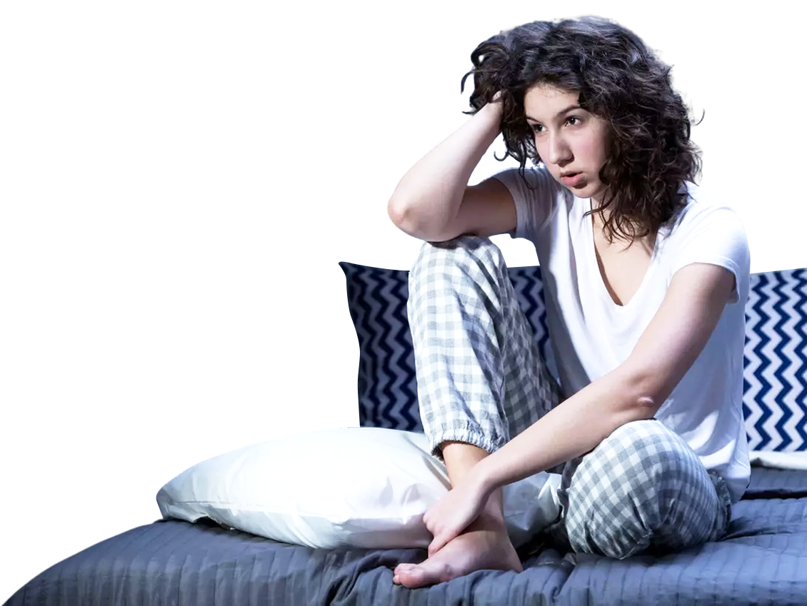 A woman wearing pyjamas sits on her bed with her legs crossed, she is running one hand through her hair and looking frustrated